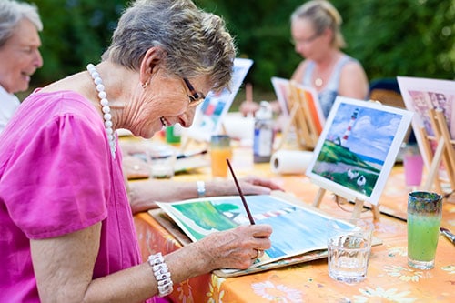 Side view of a happy senior woman smiling while paiting as a recreational activity outdoors together with a group of retired women.